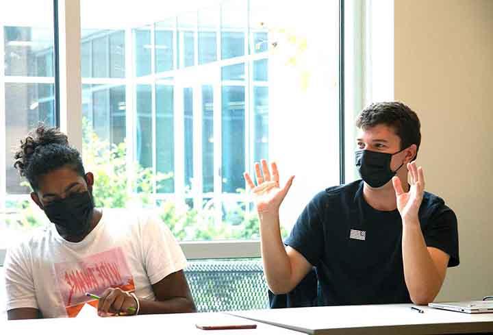 A student in a mask with both hands raised sits next to another student at a classroom table