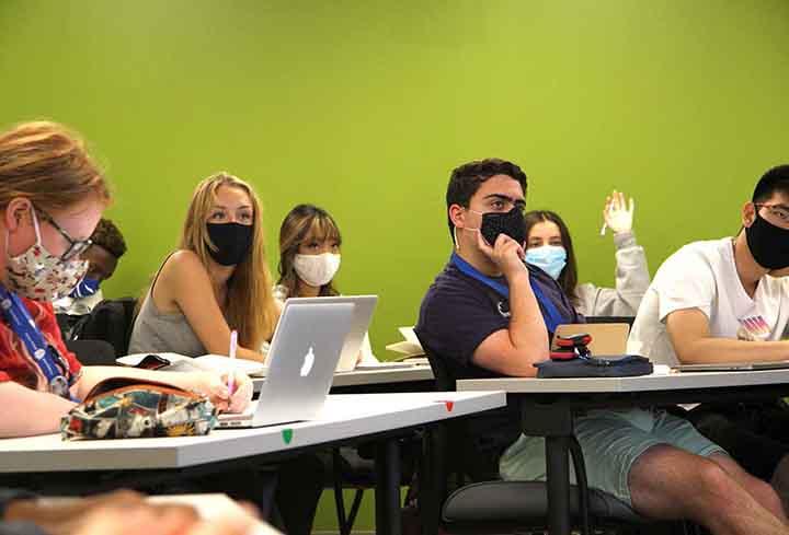 Students sitting at classroom tables in masks look ahead. One student raises her hand.