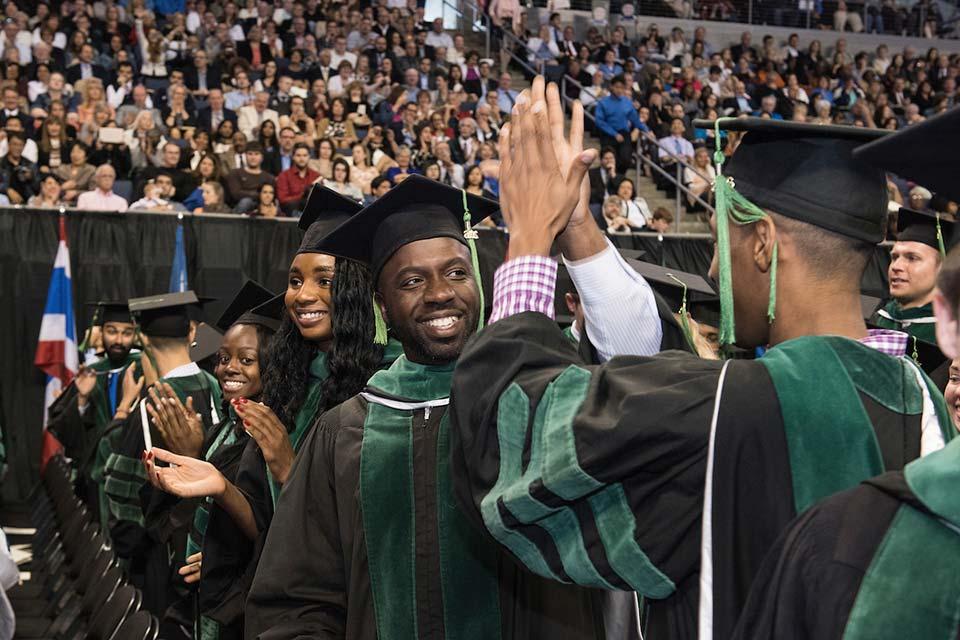 Two students wearing caps and gowns high five each other in a crowded arena.