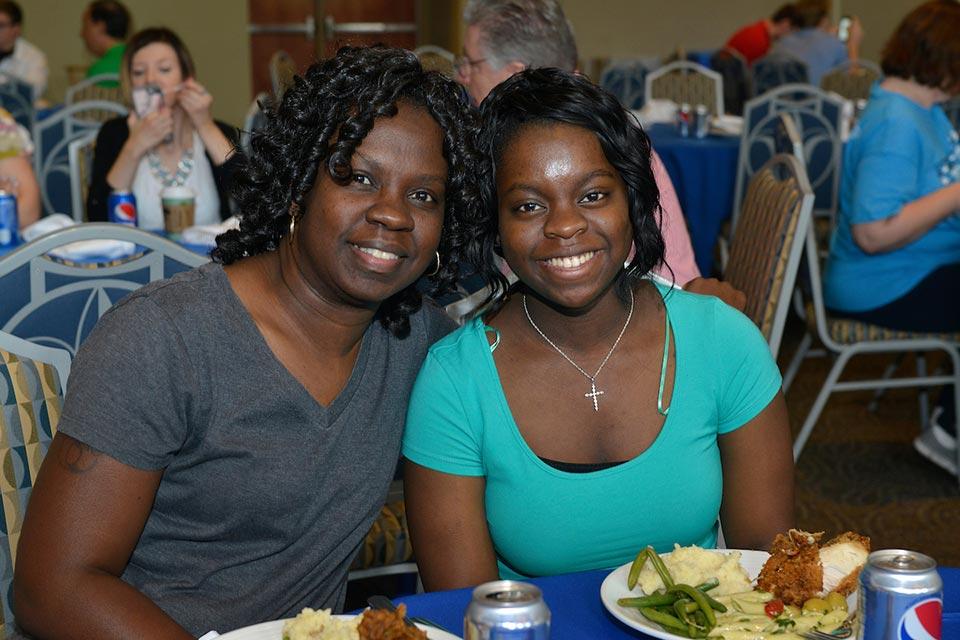 Two students pose while sitting at a dinner with plates of food and soda cans in front of them. 