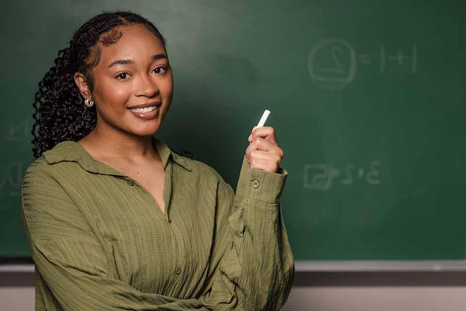 A girl poses for a photo holding chalk in front of chalk board.