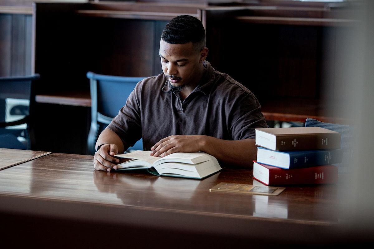 A student reads a book at a wood table in the library with a stack of books on the table next to him.