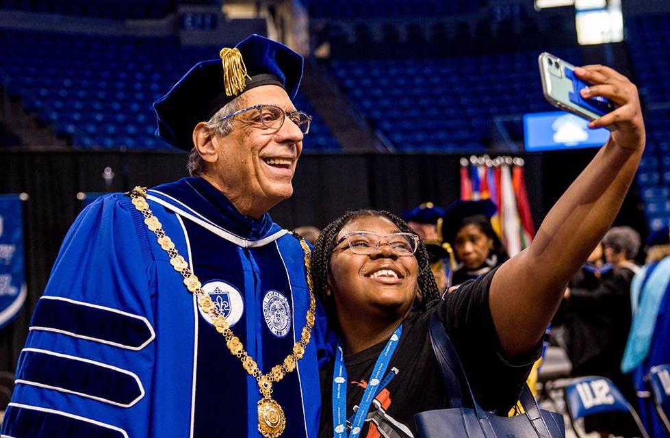 President Fred Pestello wearing a full cap and gown smiles for a selfie with a student