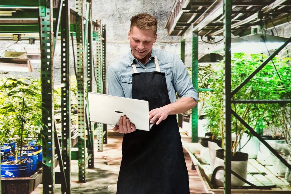 A man in an apron standing in a greenhouse holding a laptop